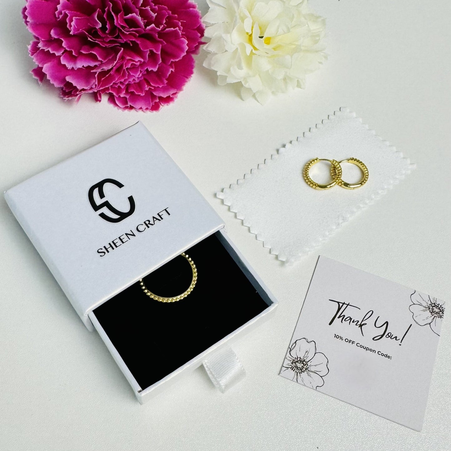 Gold jewelry set with 'Sheen Craft' box and flowers on a white background