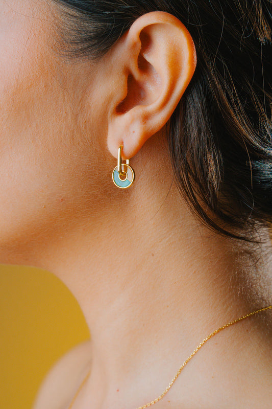 Close-up of an ear wearing a gold earring with a green Malachite and brown tiger eye gemstone, with a blurred yellow background.