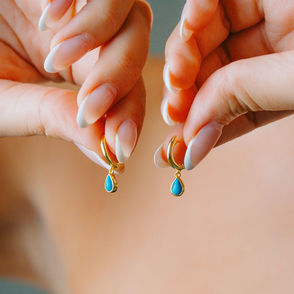 Two hands holding gold hoop earrings with turquoise drops against a neutral background
