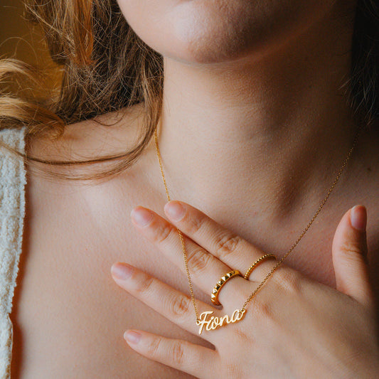 Close-up of a person wearing a gold necklace with a 'Fiona' pendant and a gold Pyramid ring.