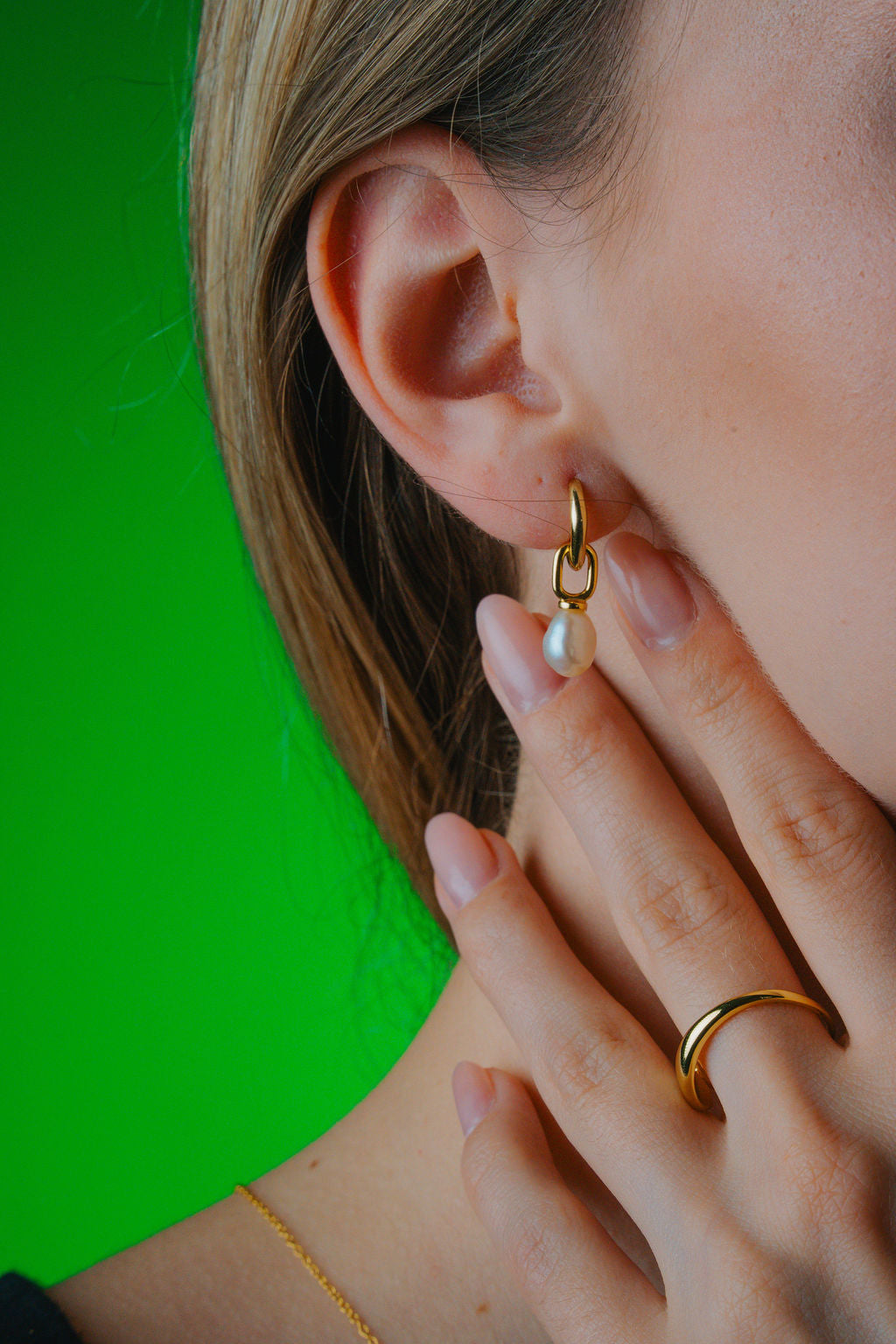Close-up of a person wearing  gold earring with a pearl and a matching ring, against a green background.