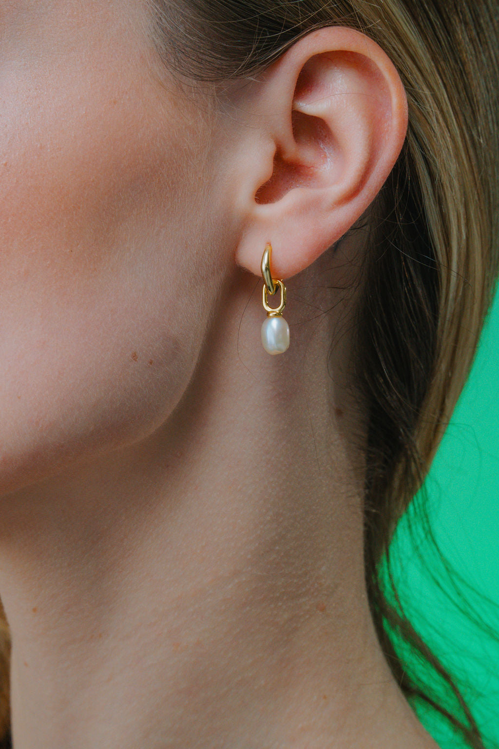 Gold earring with a pearl drop worn by a person against a green background