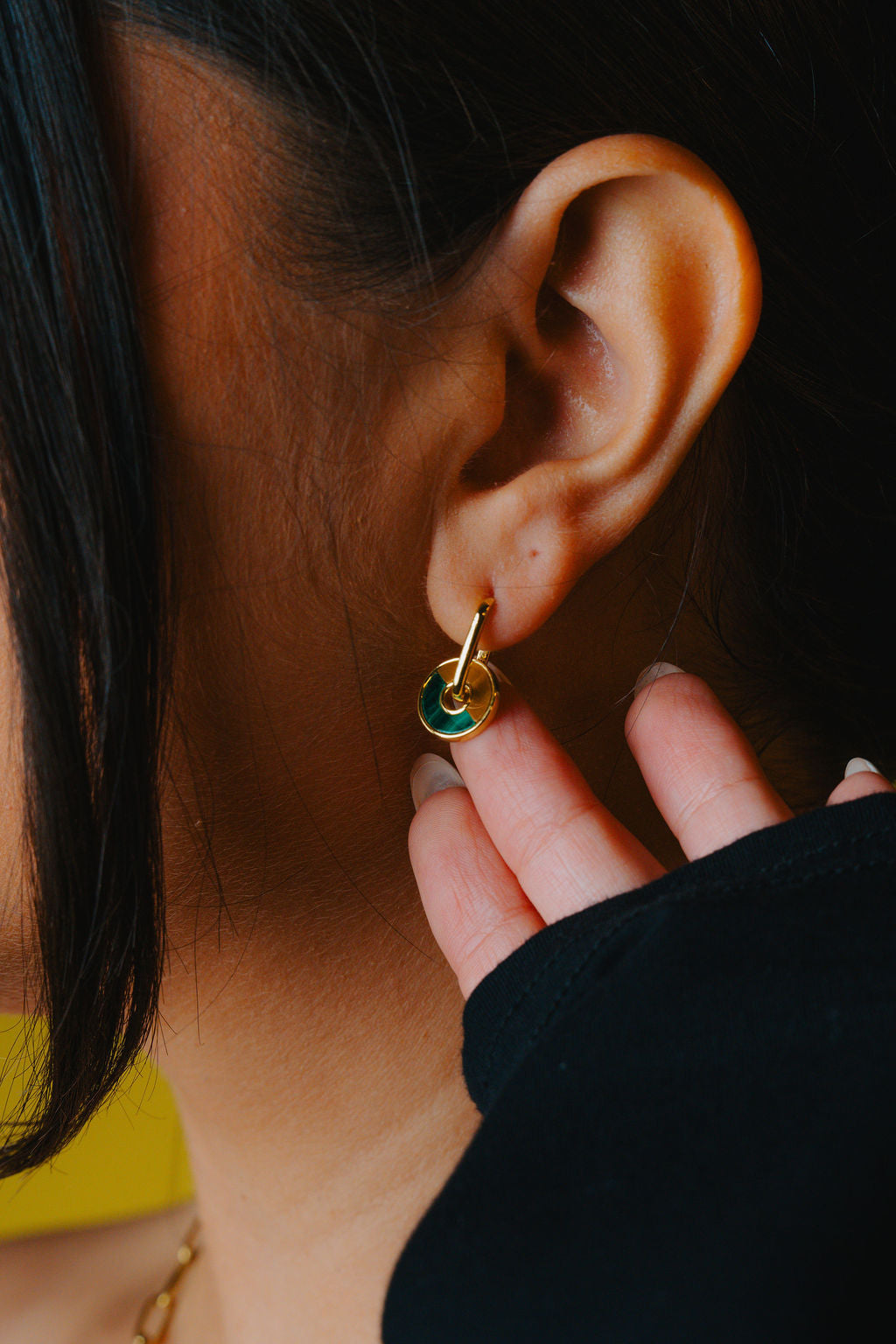 Close-up of an ear with a gold earring a green Malachite and a brown tiger eye stone hoop, hand holding the earlobe.