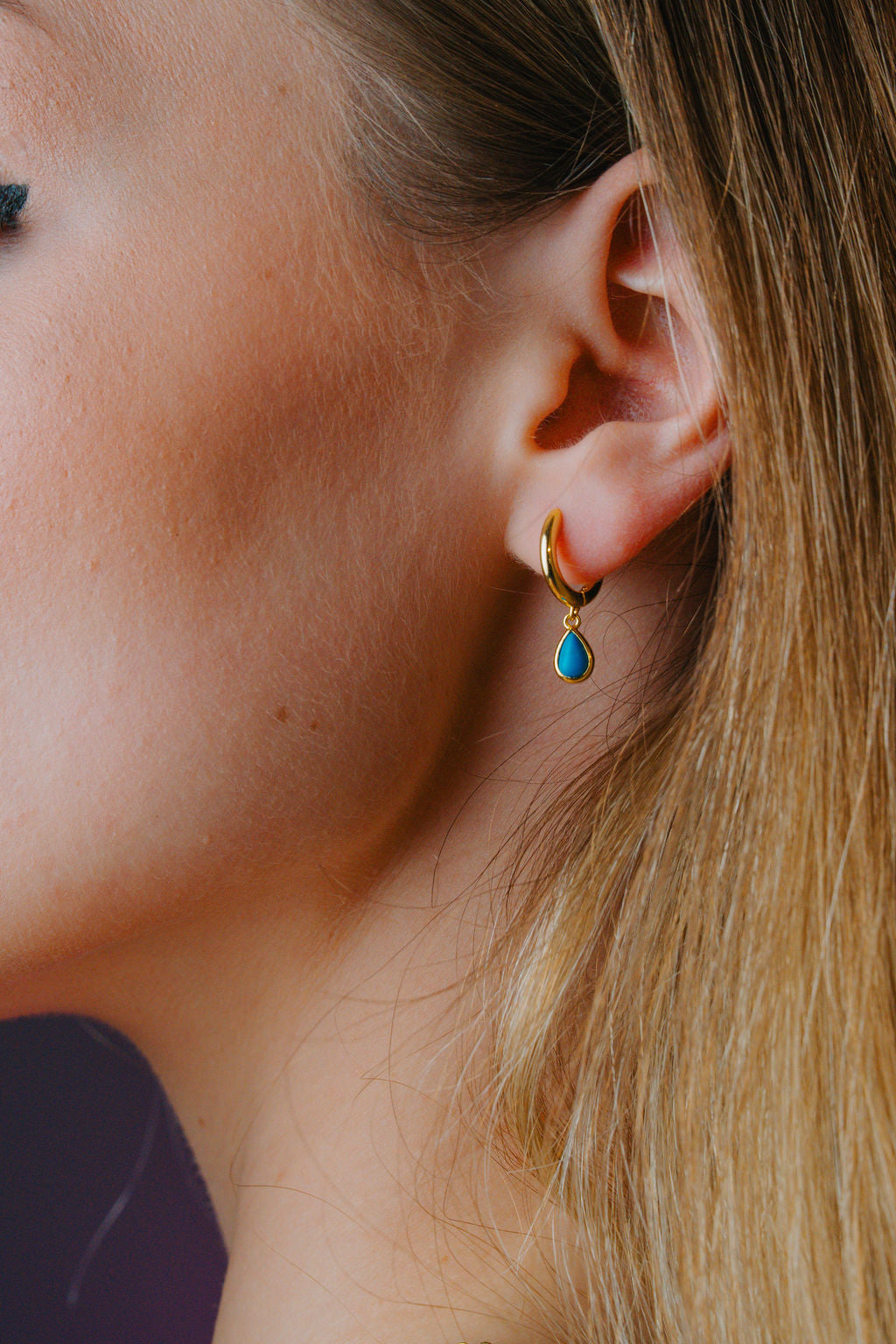 Close-up of a person wearing a gold hoop earring with a blue gemstone.