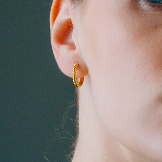 Close-up of a woman's ear wearing a gold twist huggee hoop earring against a dark background