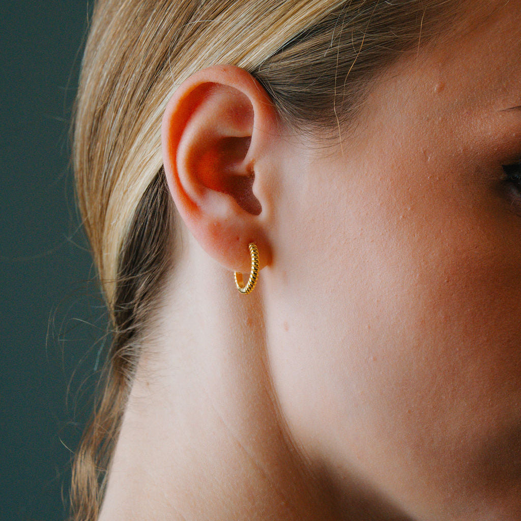 Close-up of a person wearing a Gold twist huggee hoop earring against a dark background