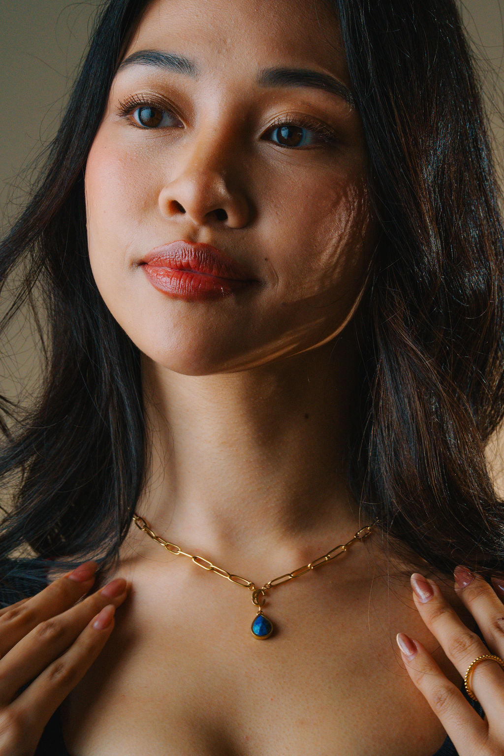 Woman wearing a gold necklace with a blue lapis lazuli pendant, with a neutral background