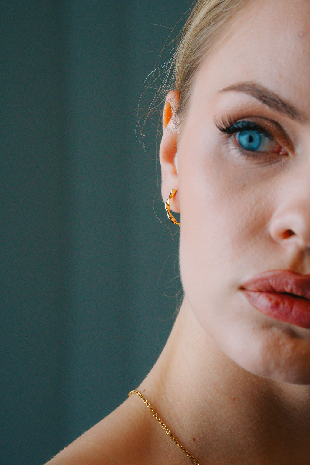 Close-up of a woman's face with Twisted Rope Large Hoop & blue eyes and gold earrings against a dark background
