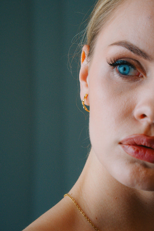Close-up of a woman's face with Twisted Rope Large Hoop & blue eyes and gold earrings against a dark background